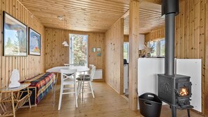 A wooden cabin interior with a central dining table, white chairs, a black stove, and a window overlooking greenery. - 3 bedroom beautiful home in Grenaa (Grenaa)