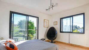 Ein modernes Schlafzimmer mit einem großen Bett, einem schwarzen Sessel und zwei Fenstern mit Blick auf Grünflächen und Gebäude.
