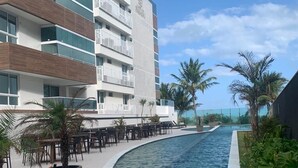 A modern hotel with a pool area, outdoor seating, and palm trees. - Apto pé na areia Ponta de Campina-PB (Cabedelo)
