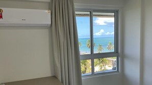 A hotel room with a bed, a window with a view of the beach, and an air conditioning unit on the wall. - Apto pé na areia Ponta de Campina-PB (Cabedelo)