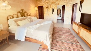 Une chambre avec un mur en pierre, un lit recouvert de draps blancs, un téléviseur posé sur un meuble en bois et un tapis à motifs.