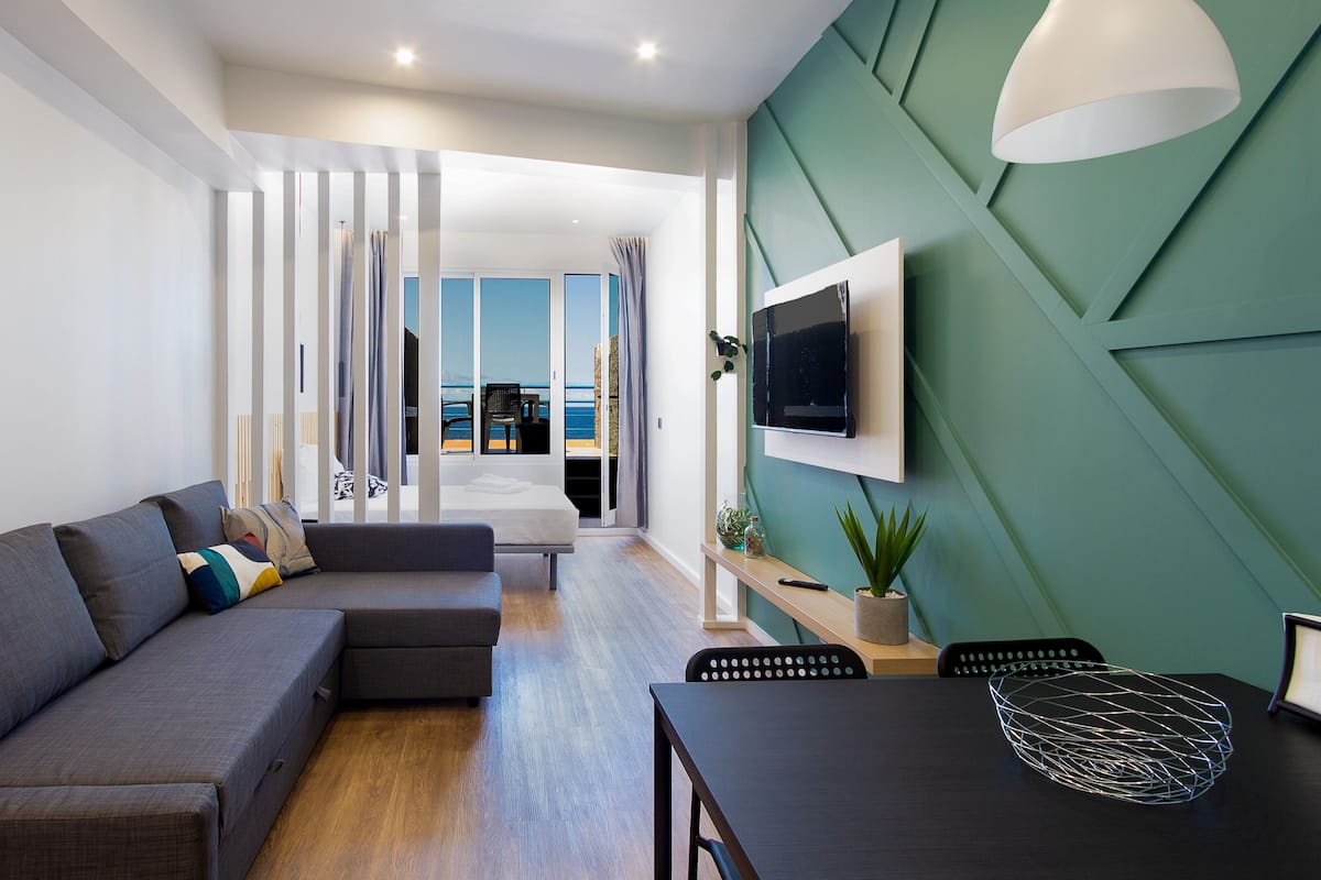Un salon moderne avec un canapé d’angle gris, une table à manger avec des chaises, un téléviseur fixé au mur et une vue sur la mer par une grande fenêtre.