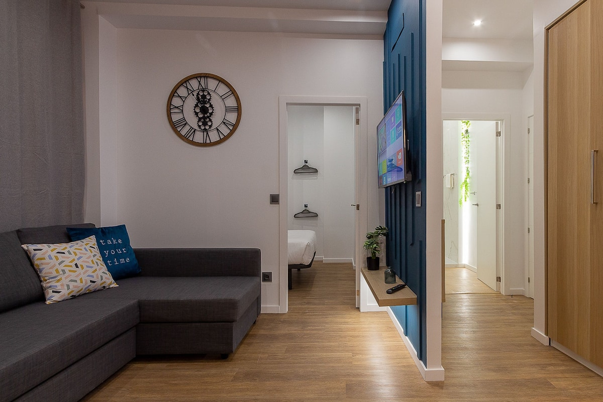 Un salon moderne avec un canapé gris, une horloge murale et un couloir menant à une chambre.