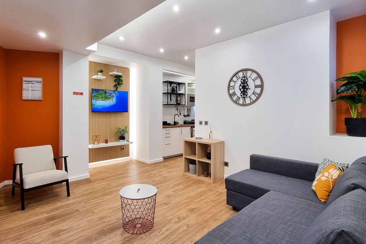 Un salon moderne avec un canapé gris, une petite table ronde et un téléviseur fixé sur un mur recouvert de panneaux en bois.