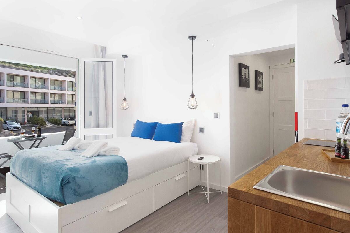 Une chambre d’hôtel moderne, équipée d’un lit, d’une petite table ronde, d’un lavabo et offrant une vue sur la rue.