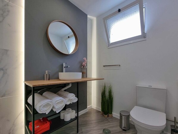 A modern bathroom with a wooden vanity, a round mirror, a toilet, and a window. - The Jolie Apartments (Porto Moniz)