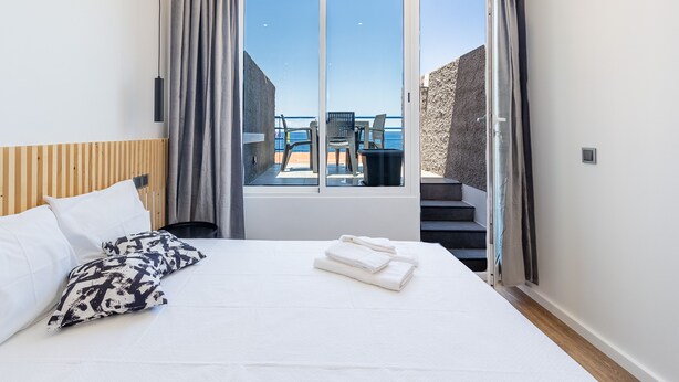 A hotel room with a bed, a view of the sea, and a balcony with chairs.