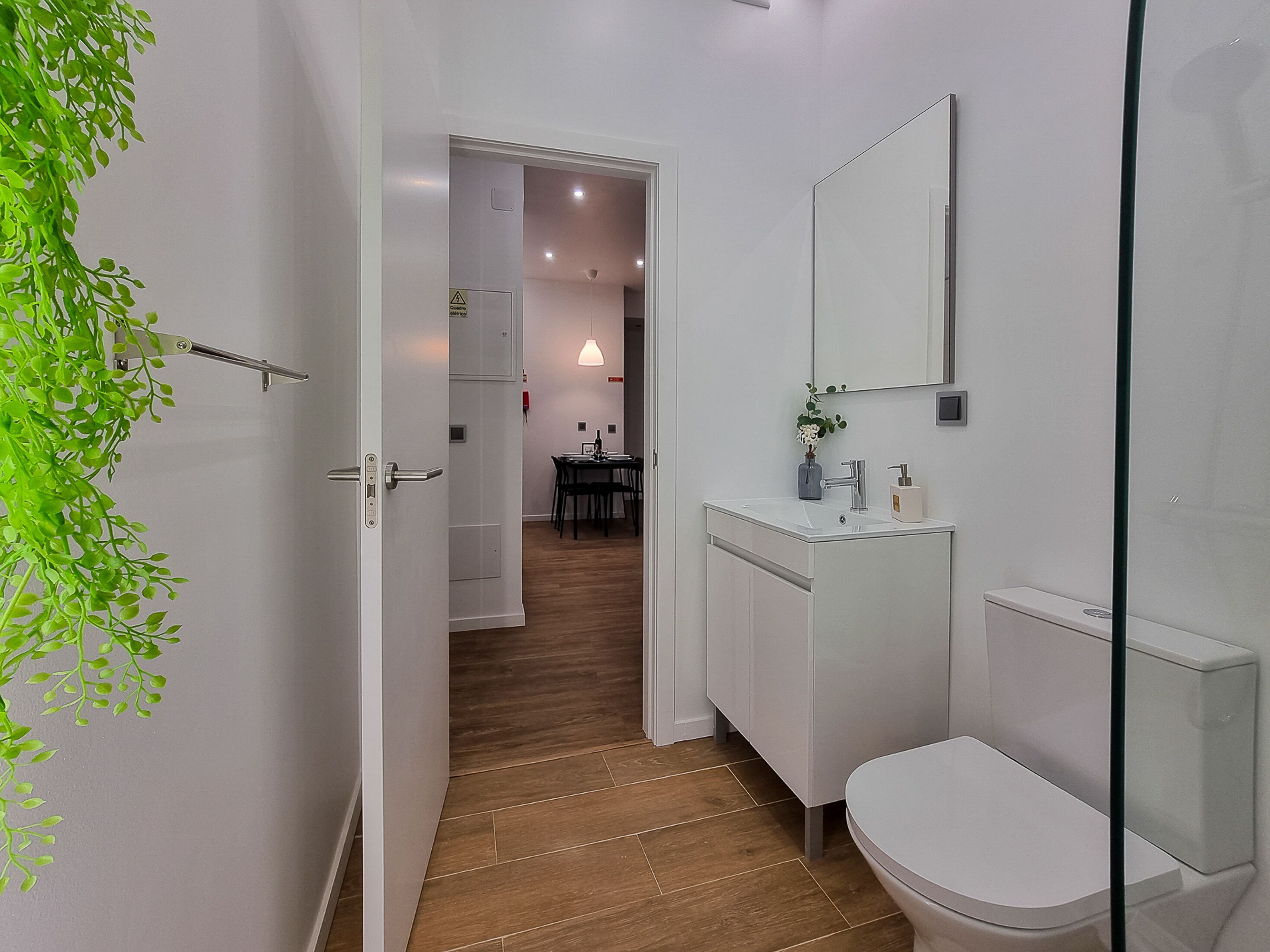 Un baño moderno con inodoro, lavabo y espejo. El suelo es de madera y hay una planta a la izquierda.