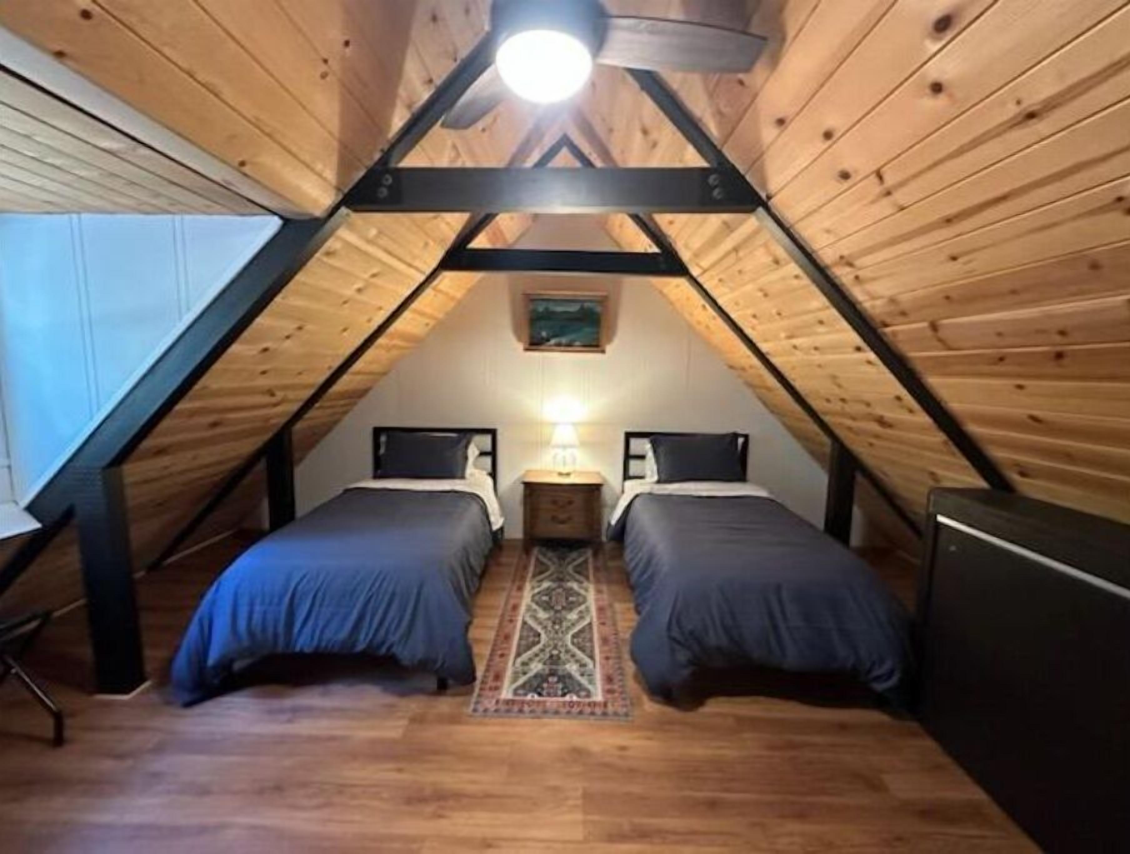 Une petite chambre mansardée de forme triangulaire, avec deux lits, une table de chevet et une lampe.