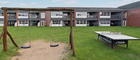 Un parque infantil con columpios y una mesa de ping pong frente a un edificio con varias ventanas y balcones.