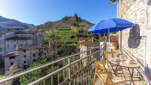 A balcony with a table and chairs, overlooking a hillside with traditional stone buildings and terraced fields.