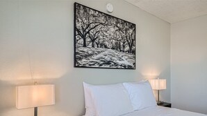 A hotel room with a bed, two bedside lamps, and a framed picture of a snowy path with trees on the wall. - Cozy Apartment with Stunning Amenities. Amazing LOCATION (Tucson)