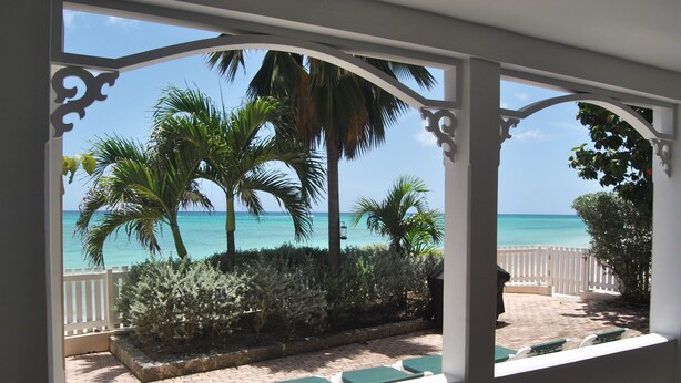 Vista a través de una ventana con marco blanco que muestra un patio con sendero de ladrillo, sillas con cojines verdes y palmeras, con vista a una playa y agua turquesa cristalina.