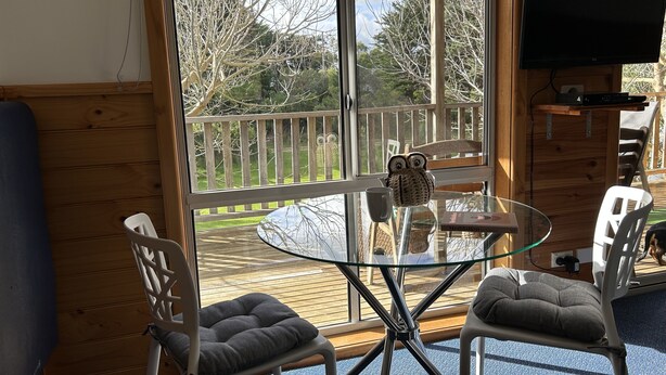 A room with a glass table, two cushioned chairs, a wooden deck outside, and a TV mounted on the wall.