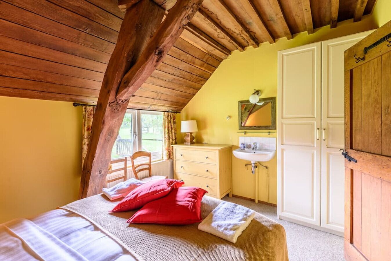 Een slaapkamer met een schuine balkenlaag, een bed met rode en witte kussens, een houten stoel, een witte ladekast, een wastafel en een kledingkast.