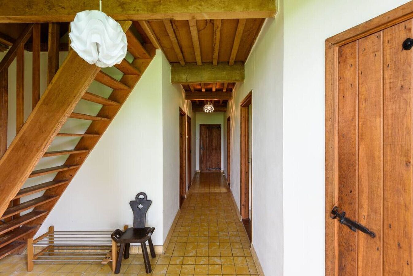 Eine Holztreppe führt zu einem Flur mit Fliesenboden, einer Tür rechts und einem Stuhl im Vordergrund.