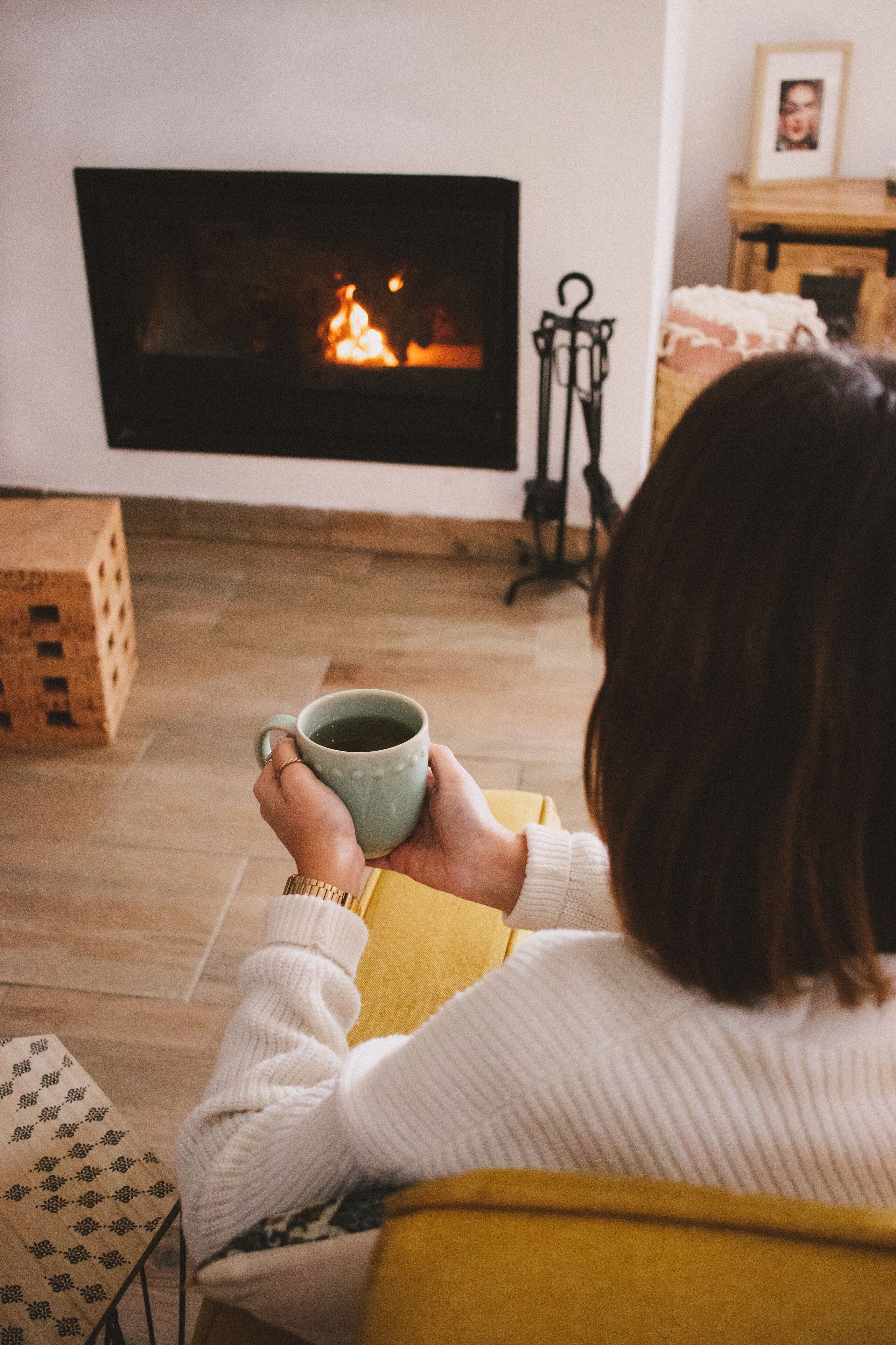 A person holding a mug, sitting in a room with a fireplace, wooden flooring, and a framed picture on the wall.