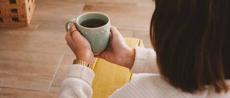 A person holding a mug, sitting in a room with a fireplace, wooden flooring, and a framed picture on the wall.