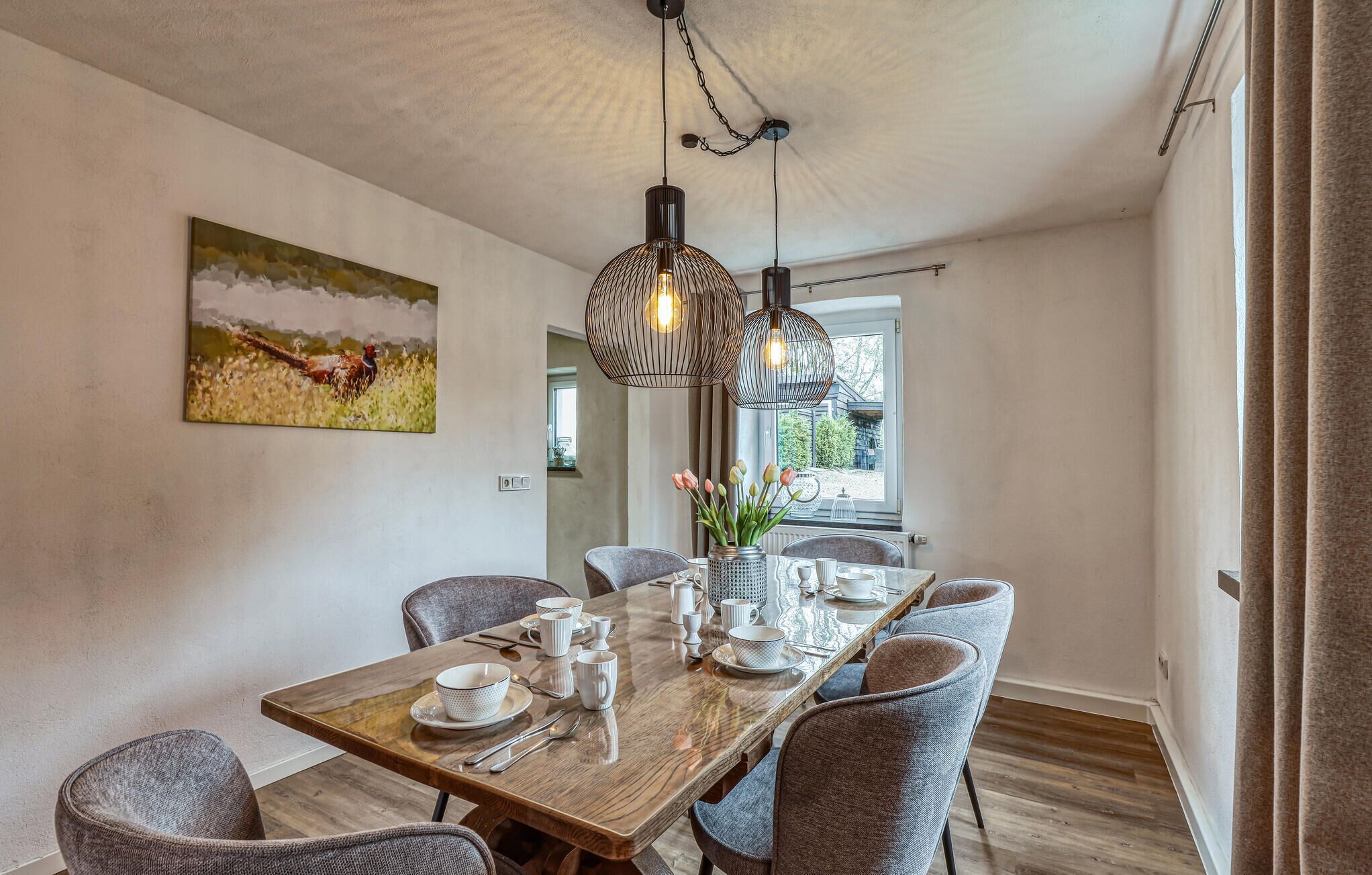 A dining area with a wooden table set for four, surrounded by grey upholstered chairs. There are pendant lights hanging above the table, and a painting of a rooster on the wall.