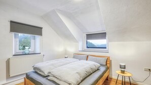 A bedroom with a sloped ceiling, a bed with grey bedding, two windows with blinds, a radiator, and a small bedside table with a lamp. - Beautiful home in Nimsreuland with WiFi (Nimsreuland)