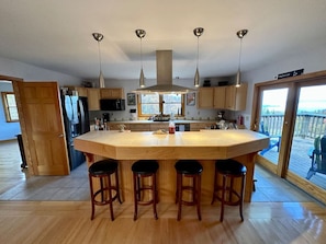A modern kitchen with a large island, bar stools, pendant lights, and a view through the glass door.