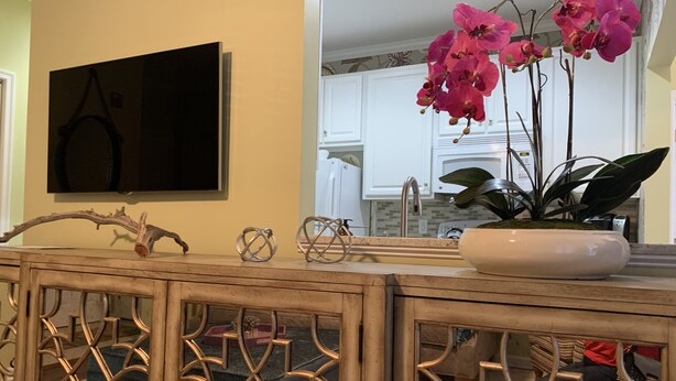 Una consola de madera con detalles metálicos decorativos, un florero blanco con orquídeas rosadas y un televisor de pantalla plana montado en la pared.
