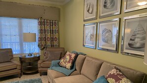 Sala de estar con un sofá color beige, cojines estampados, un plaid azul y una mesa de centro con objetos decorativos. En la pared hay cuadros enmarcados y una ventana con persianas.