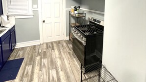 Une cuisine compacte équipée d’une cuisinière à gaz, d’un four et d’une hotte en acier inoxydable. On y trouve une porte blanche et une fenêtre avec des stores.