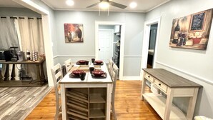 Un coin repas avec une table blanche dressée pour quatre personnes, un ventilateur de plafond et un tableau accroché au mur.