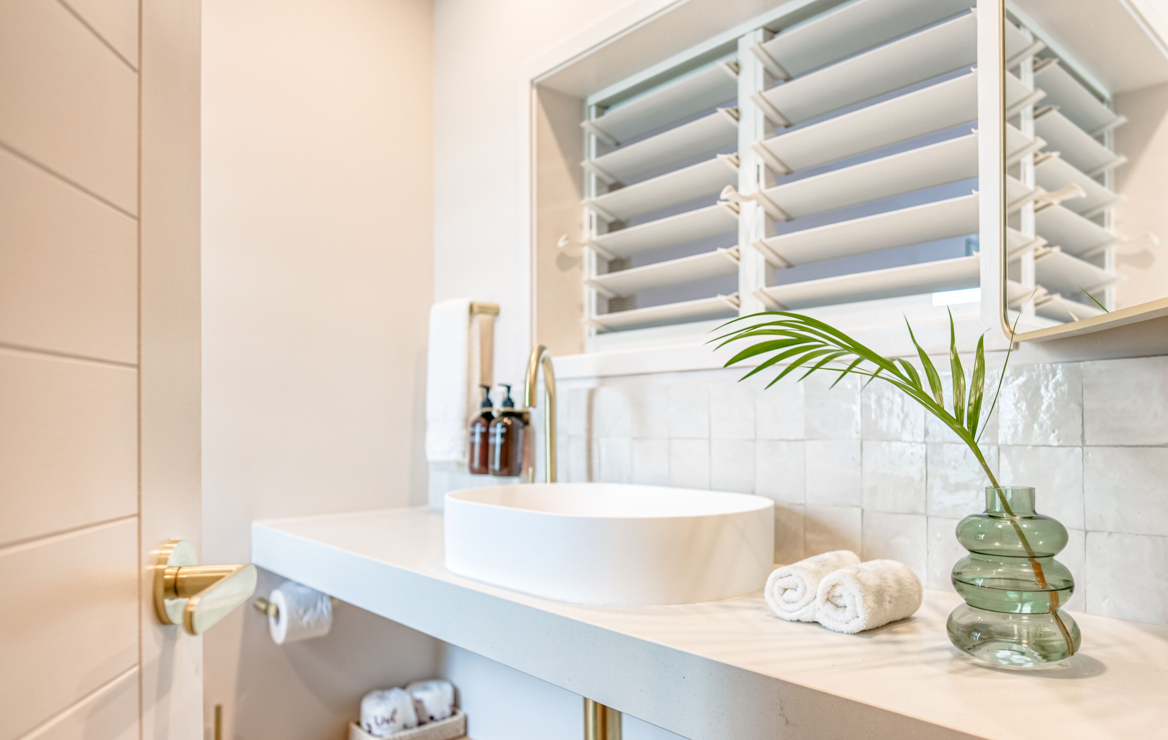 A bathroom with a white sink, a mirror, and a window with blinds. There are towels and a vase with a plant on the counter.