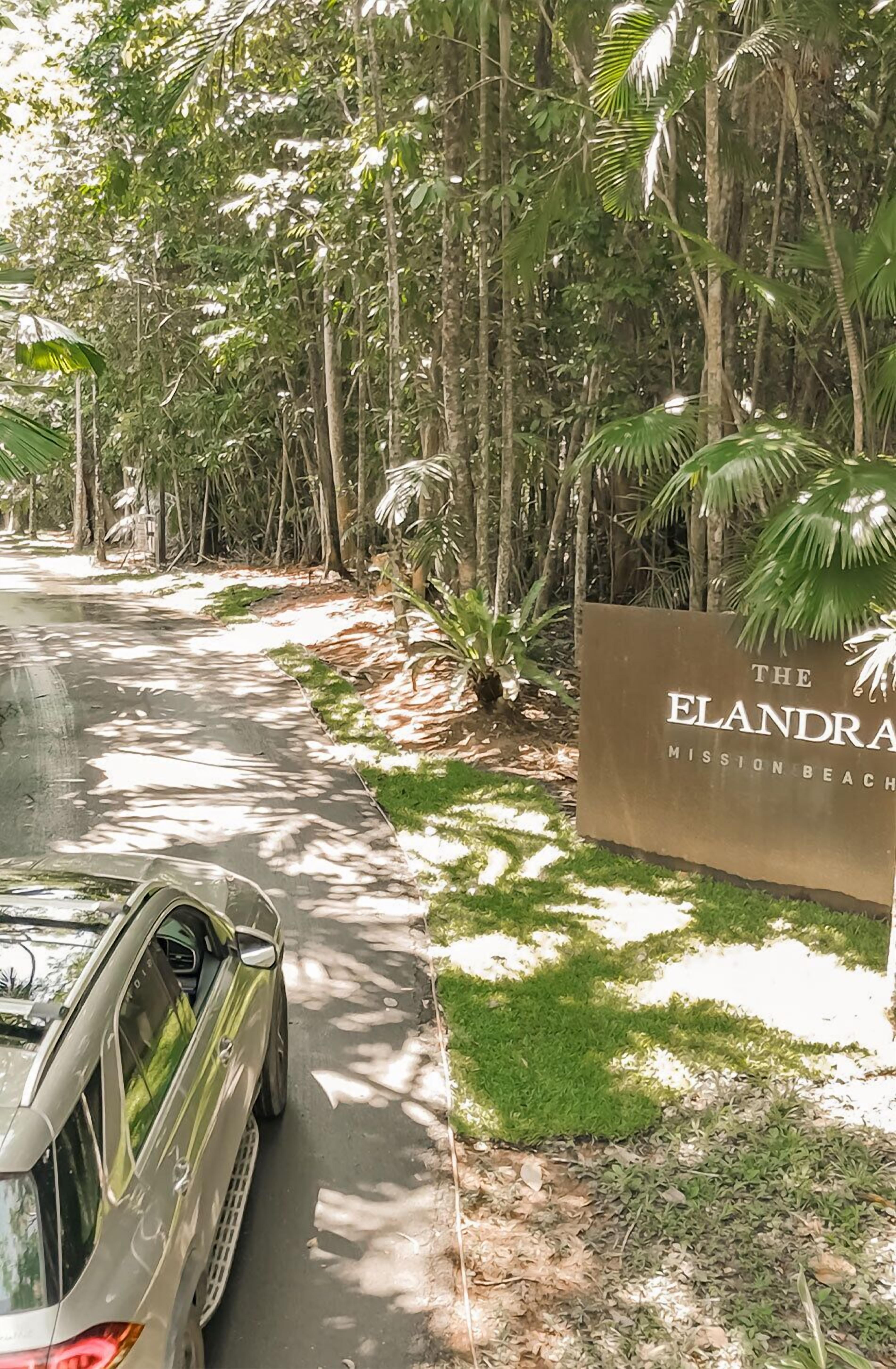 Un camino pavimentado con un coche estacionado, rodeado de abundante vegetación y un cartel de The Elandra Mission Beach.