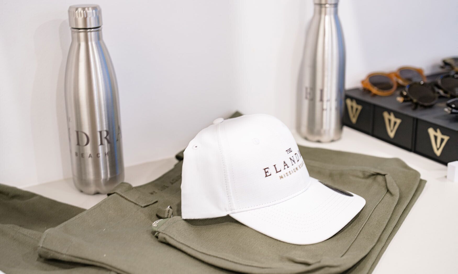 A white cap with "ELAND" text, a green jacket, and two stainless steel bottles on a white surface.