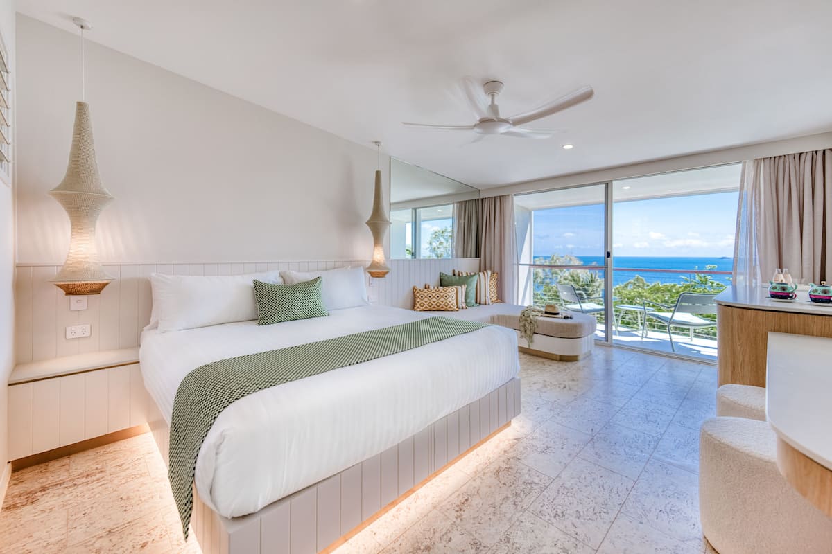 A modern hotel room with a large bed, a balcony with a view of the ocean, and a ceiling fan.