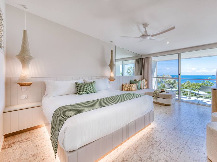 A modern hotel room with a large bed, a balcony with a view of the ocean, and a ceiling fan.