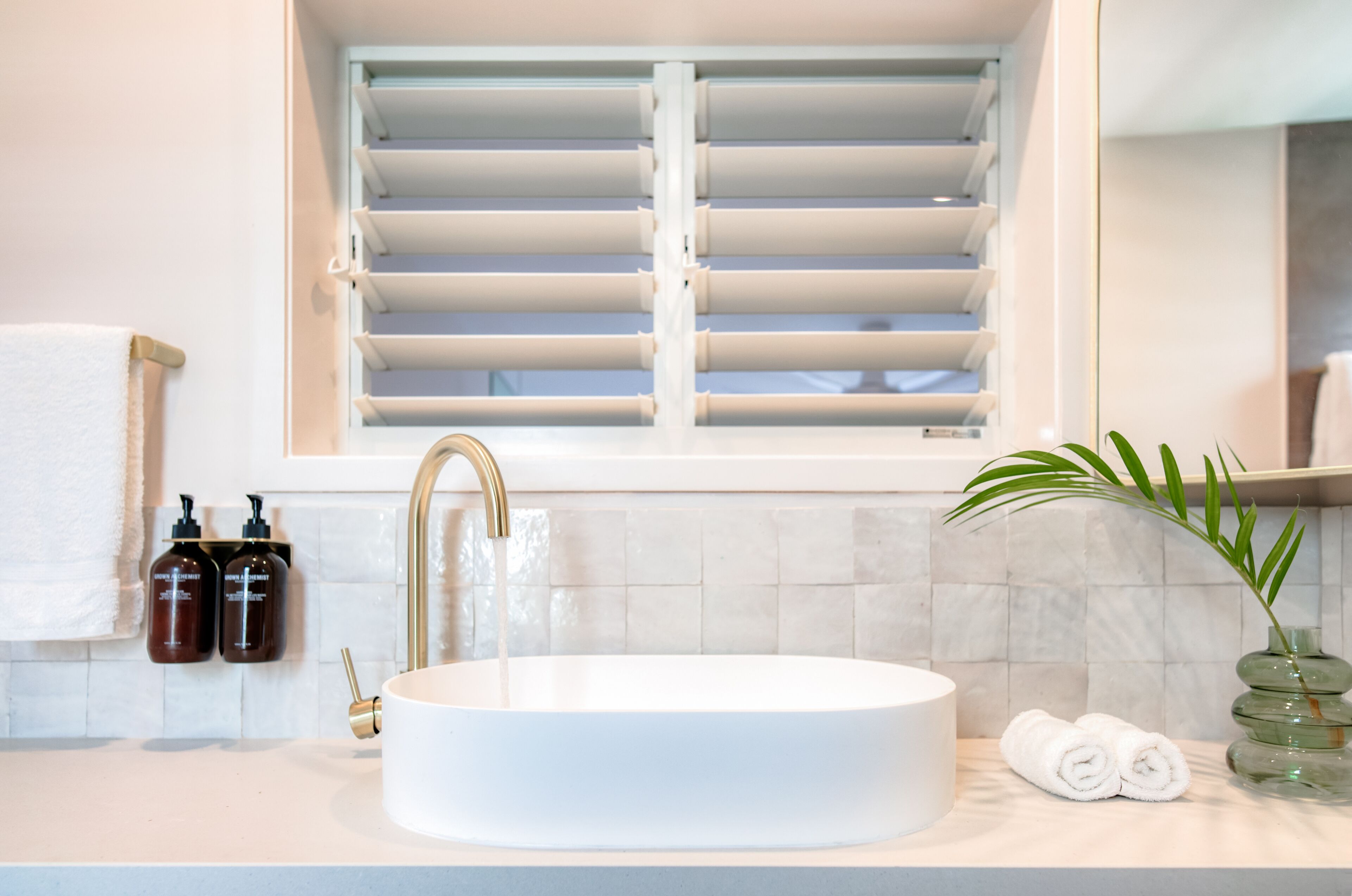 A bathroom with a white sink, a gold faucet, and a window with white blinds. There are two bottles of shampoo and a rolled towel on the counter.