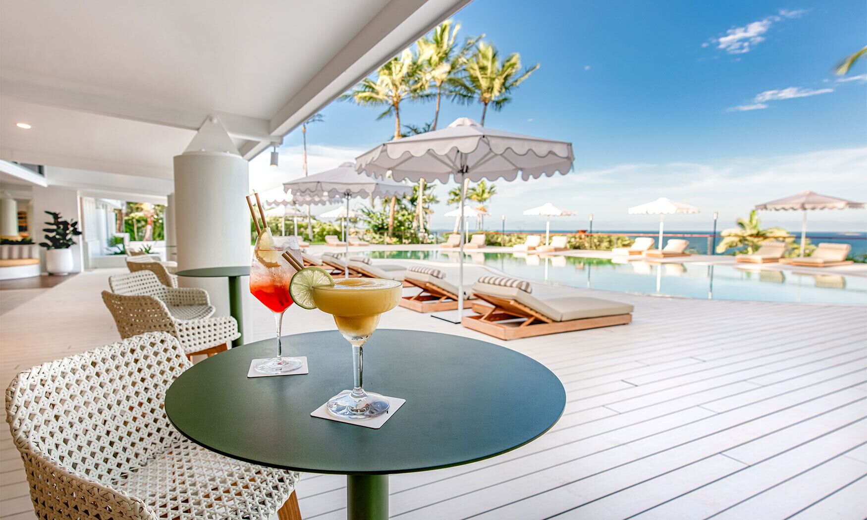Een zwembadterras met ligbedden en parasols, een tafel met drankjes, en uitzicht op de oceaan.