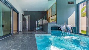 A modern indoor swimming pool with a waterfall feature, surrounded by glass walls and a dark tiled floor. - Amazing home in Fazana with sauna (Fazana)