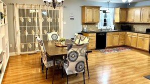A dining area with a wooden table and chairs, a chandelier, and a kitchen with wooden cabinets and a microwave.
