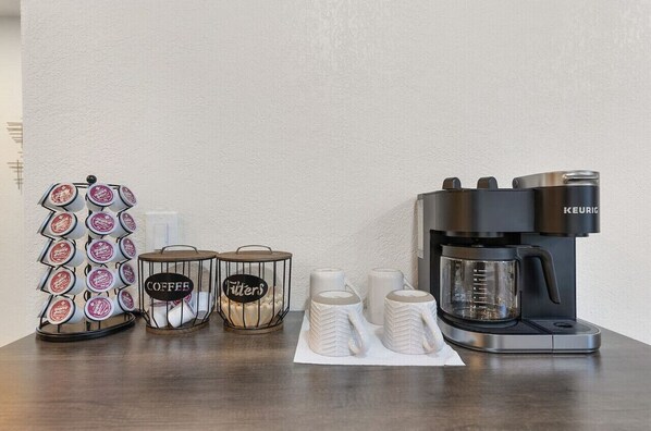 En kaffestation med en Keurig-kaffemaskin, en behållare för kaffekapslar, två glasburkar märkta med "Kaffe" och "Filter", och tre vita muggar på ett bord.