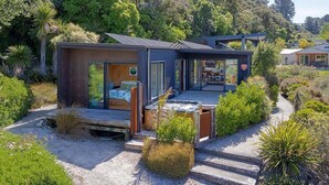 Een modern, compact huis met een terras, omgeven door weelderige begroeiing en een grindpad.