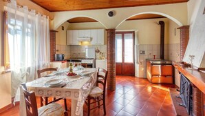 Comedor con una mesa dispuesta para cuatro, cocina con armarios blancos, un pilar de ladrillo y un techo de paneles de madera.
