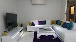 Un salon moderne avec un canapé blanc, un téléviseur à écran plat, une table basse en verre et un tapis violet.
