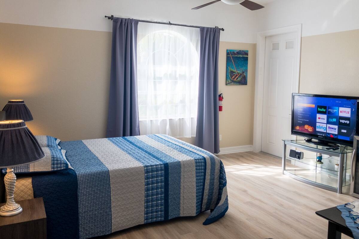 Una habitación de hotel con cama, televisión, ventilador de techo y ventana con cortinas.