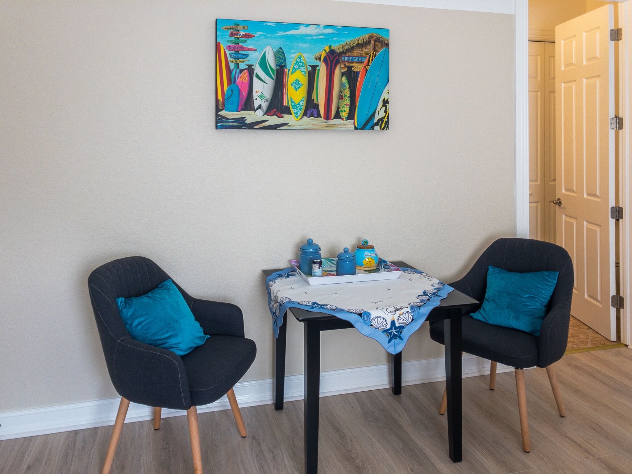 A room with two black chairs, a small table with a blue tablecloth, and a colorful painting on the wall.