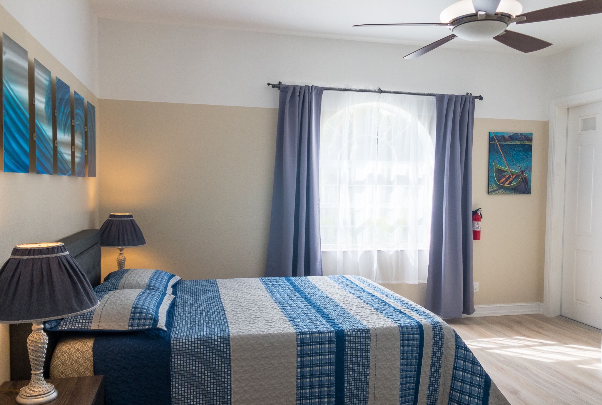 Een slaapkamer met een bed, twee bedlampen, een plafondventilator, een raam met gordijnen, en een brandblusser aan de muur.