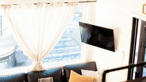 Una sala de estar acogedora con techo de madera, ventilador de techo, televisor de pantalla plana montado en la pared, sofá con cojines y una ventana con cortinas.