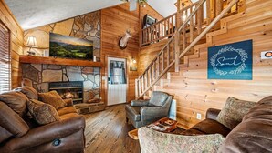 Interior acogedor de una cabaña con chimenea, sofás de cuero, una escalera de madera y una obra de arte enmarcada en la pared.