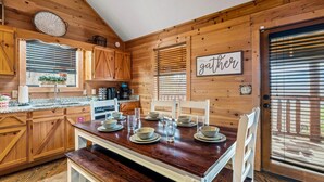 Cocina de cabaña de madera con una mesa de comedor preparada para cuatro comensales, armarios de madera, un reloj y un letrero que dice 'reunirse' en la pared.