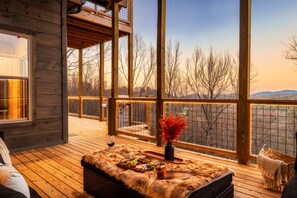 Une terrasse en bois offrant une vue sur les arbres et les montagnes, une porte vitrée et une table agrémentée d’un vase et de fruits.