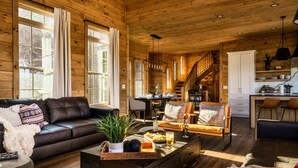 A spacious living room with a leather sofa, a coffee table with a tray of snacks, and a staircase leading to a kitchen area. - New to VRBO!  Theater Seating, Pool Table, Gaming Station, Bunk Rm, Hot Tub (Gatlinburg)